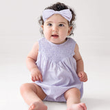A baby with curly hair and a light purple headband wears the Magnolia Sleeveless Romper by Mabel and Honey, sitting on a white floor and gazing upward with a curious expression.