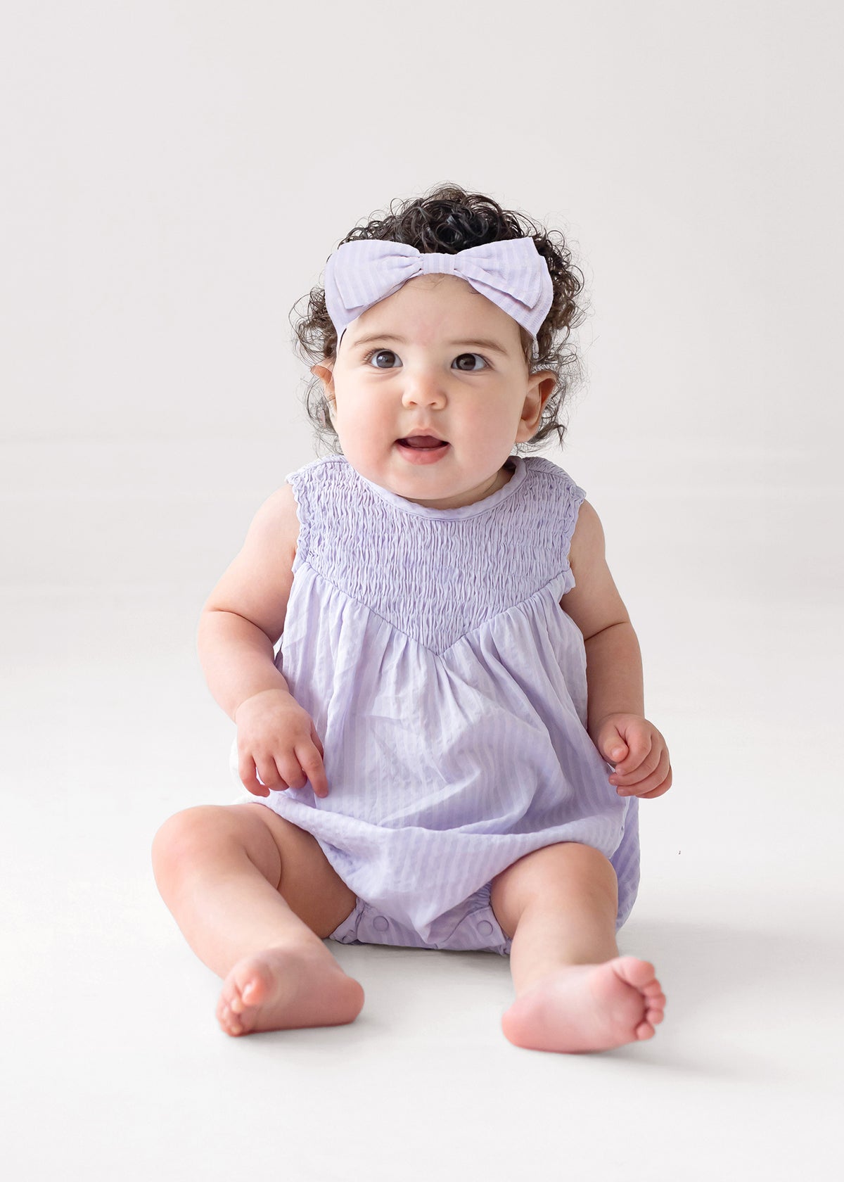 A baby with curly hair and a light purple headband wears the Magnolia Sleeveless Romper by Mabel and Honey, sitting on a white floor and gazing upward with a curious expression.