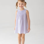 A young girl with blonde pigtails is smiling against a white background, wearing the Mabel and Honey Magnolia Sleeveless Dress in lavender paired with beige sandals.