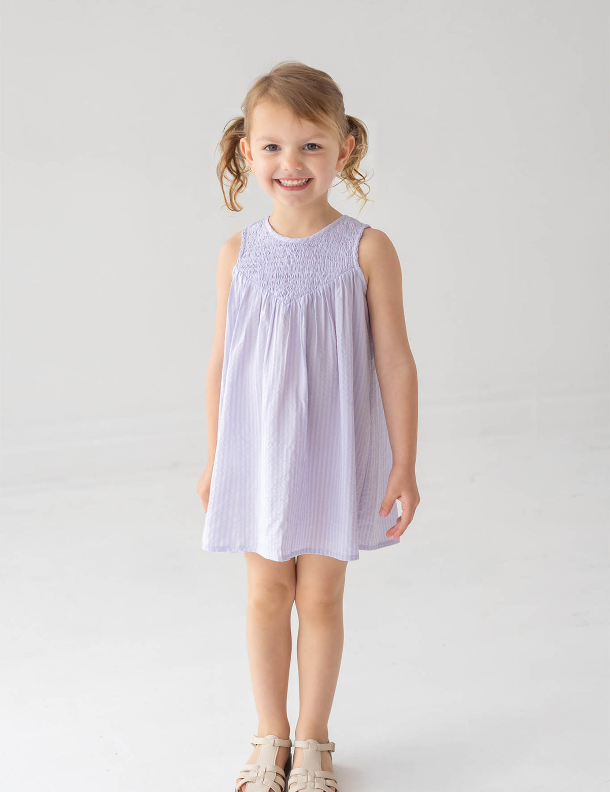 A young girl with blonde pigtails is smiling against a white background, wearing the Mabel and Honey Magnolia Sleeveless Dress in lavender paired with beige sandals.