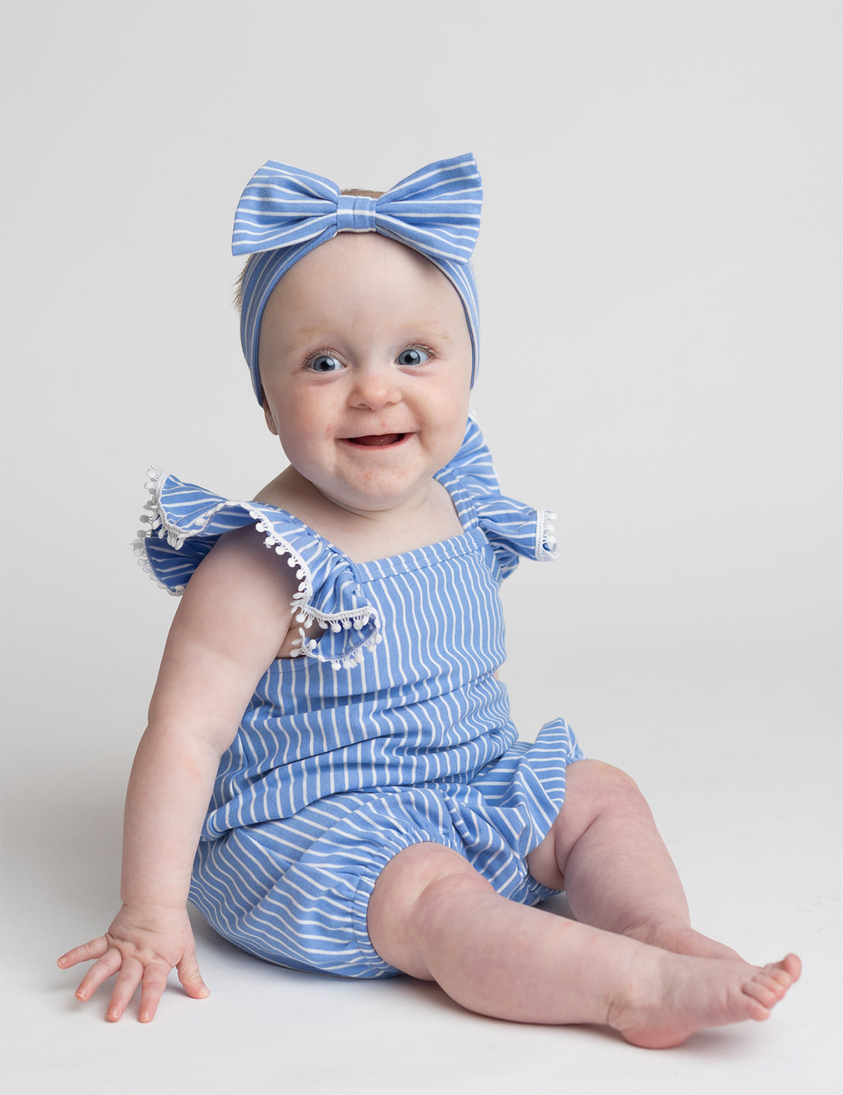 A smiling baby sits on the floor against a plain light background, wearing the Mabel and Honey Piper Ruffled Tank Romper in light blue with ruffled shoulder straps and a matching large-bow headband.