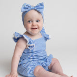A smiling baby sits on the floor against a plain light background, wearing the Mabel and Honey Piper Ruffled Tank Romper in light blue with ruffled shoulder straps and a matching large-bow headband.