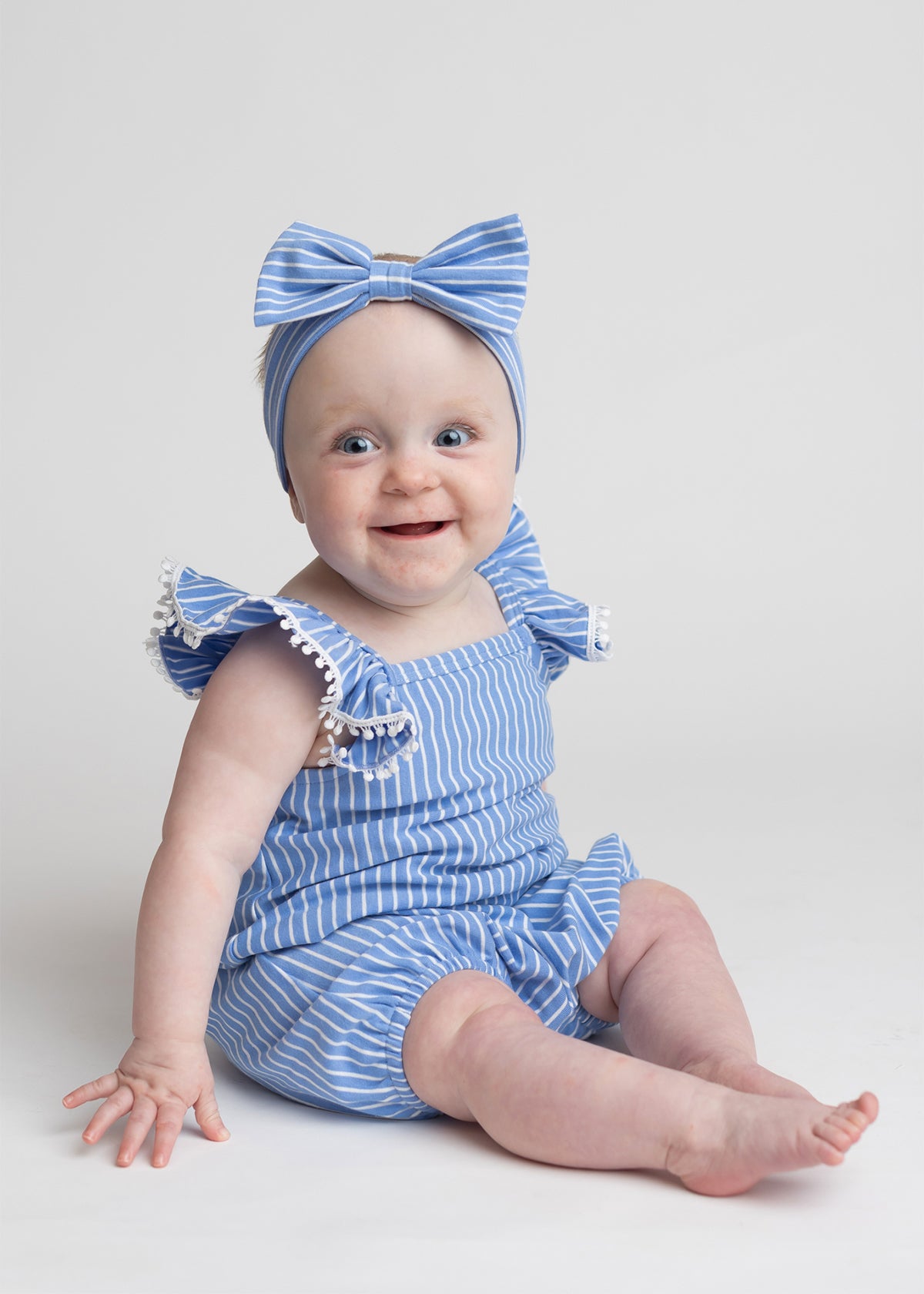 A smiling baby sits on the floor against a plain light background, wearing the Mabel and Honey Piper Ruffled Tank Romper in light blue with ruffled shoulder straps and a matching large-bow headband.