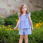 A young girl with light brown hair wears the Mabel and Honey Piper Ruffled Tank Two Piece Set, featuring blue and white stripes, flutter sleeves, and matching shorts, standing on a path by green bushes with yellow flowers and a stone wall.