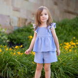 A young girl with light brown hair wears the Mabel and Honey Piper Ruffled Tank Two Piece Set, featuring blue and white stripes, flutter sleeves, and matching shorts, standing on a path by green bushes with yellow flowers and a stone wall.