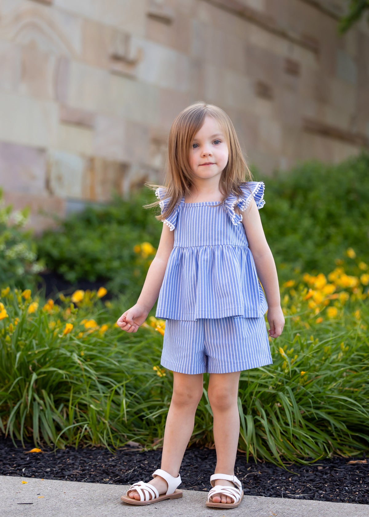 A young girl with light brown hair wears the Mabel and Honey Piper Ruffled Tank Two Piece Set, featuring blue and white stripes, flutter sleeves, and matching shorts, standing on a path by green bushes with yellow flowers and a stone wall.