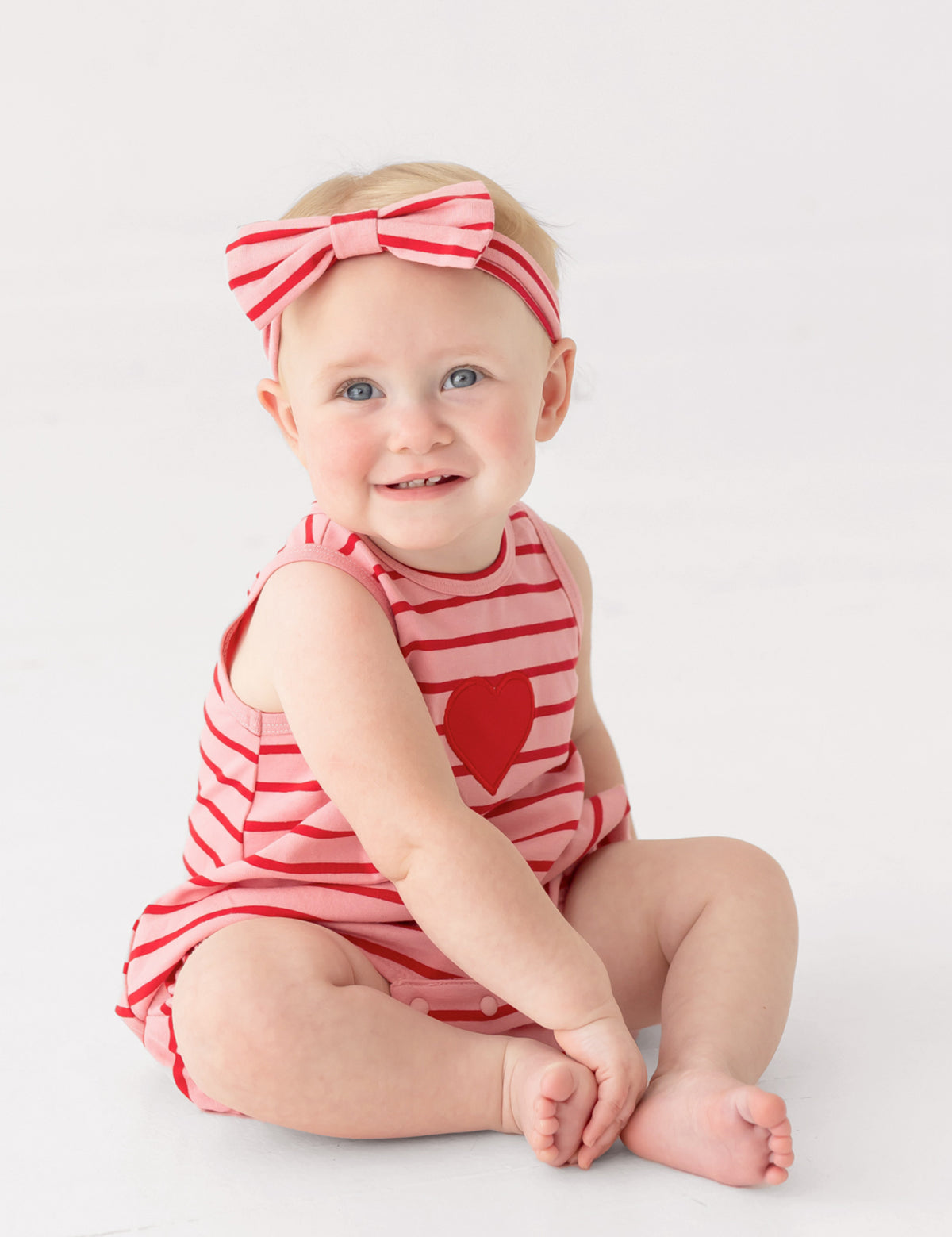 A smiling baby sits barefoot on a white floor wearing the Mabel and Honey Sugar Sweet Sleeveless Romper in red and white stripes with a heart applique, paired with a matching striped bow headband.