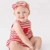 A smiling baby sits barefoot on a white floor wearing the Mabel and Honey Sugar Sweet Sleeveless Romper in red and white stripes with a heart applique, paired with a matching striped bow headband.