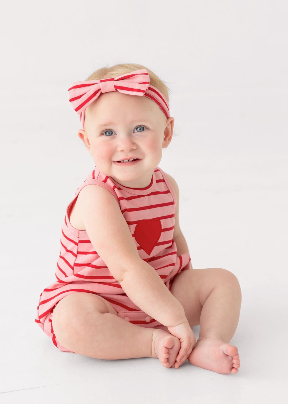 A smiling baby sits barefoot on a white floor wearing the Mabel and Honey Sugar Sweet Sleeveless Romper in red and white stripes with a heart applique, paired with a matching striped bow headband.