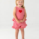 A young blonde-haired child smiles barefoot, wearing the Sugar Sweet Sleeveless Two Piece Set by Mabel and Honey—ruffled shorts and a red heart applique on a red and white striped outfit—standing on a white background.
