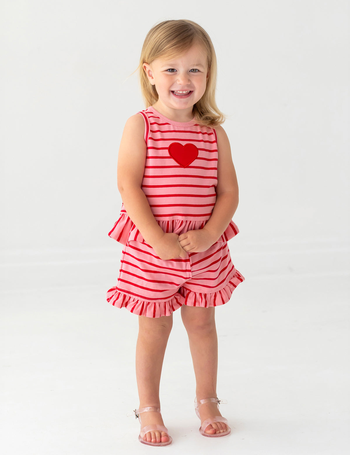 A young blonde-haired child smiles barefoot, wearing the Sugar Sweet Sleeveless Two Piece Set by Mabel and Honey—ruffled shorts and a red heart applique on a red and white striped outfit—standing on a white background.