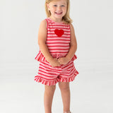 A young blonde-haired child smiles barefoot, wearing the Sugar Sweet Sleeveless Two Piece Set by Mabel and Honey—ruffled shorts and a red heart applique on a red and white striped outfit—standing on a white background.