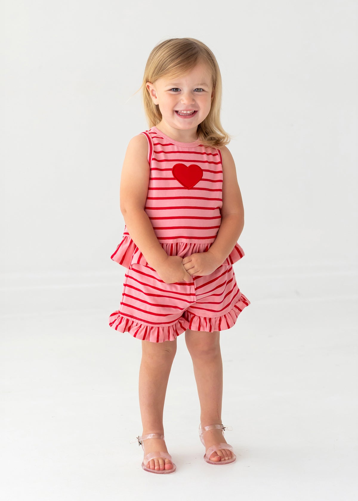 A young blonde-haired child smiles barefoot, wearing the Sugar Sweet Sleeveless Two Piece Set by Mabel and Honey—ruffled shorts and a red heart applique on a red and white striped outfit—standing on a white background.
