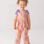 A young child smiles, wearing the Mabel and Honey Petal Power Tank Sleeve with Knot Detail Two Piece Set—featuring a colorful geometric floral tunic and matching wide-leg pants—paired with white sandals against a plain light background.