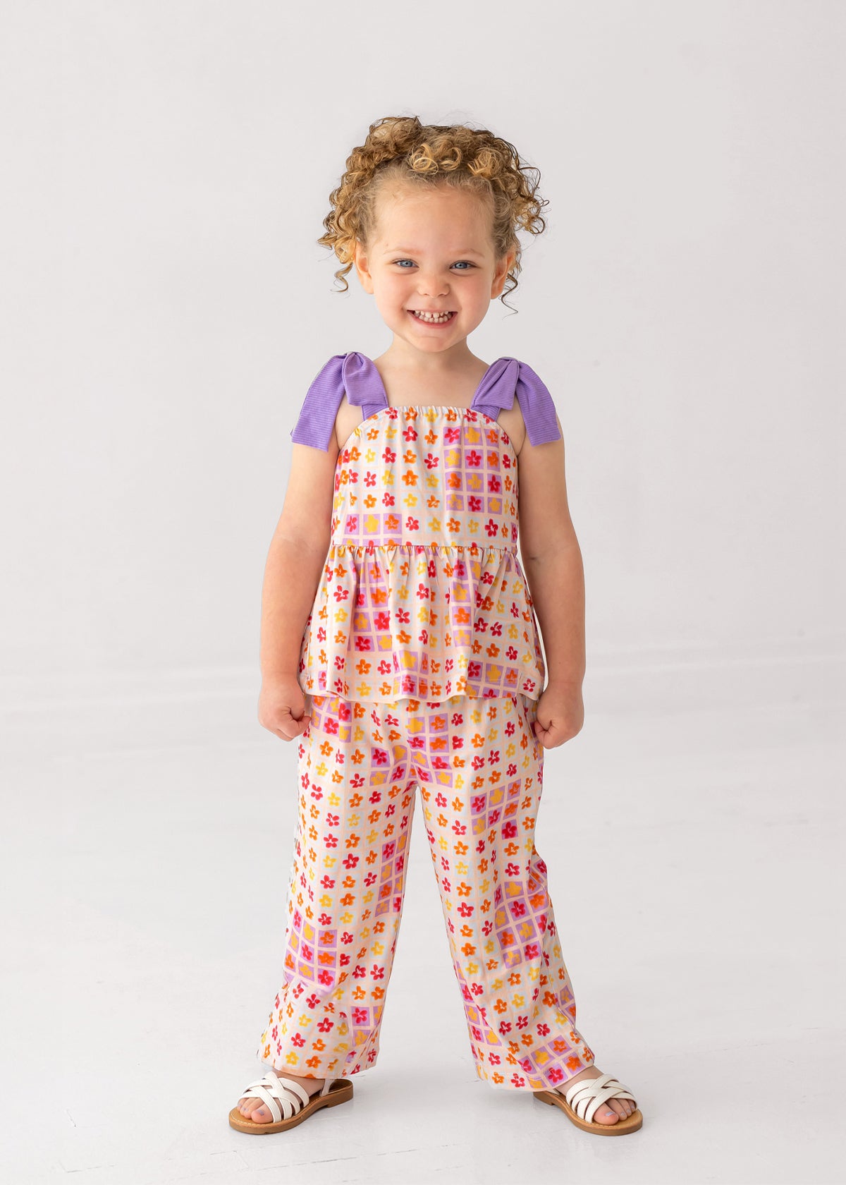 A young child smiles, wearing the Mabel and Honey Petal Power Tank Sleeve with Knot Detail Two Piece Set—featuring a colorful geometric floral tunic and matching wide-leg pants—paired with white sandals against a plain light background.