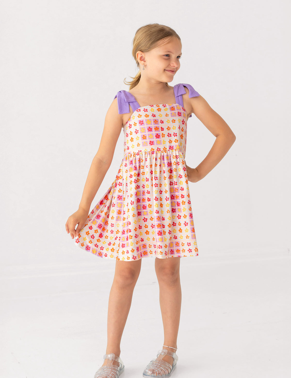 A young girl smiles in the Mabel and Honey Petal Power Tank Sleeve with Knot Detail Dress, featuring a colorful geometric print, purple shoulder knots, and an A-line skirt. She stands on a white background, wearing clear sandals.