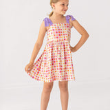 A young girl smiles in the Mabel and Honey Petal Power Tank Sleeve with Knot Detail Dress, featuring a colorful geometric print, purple shoulder knots, and an A-line skirt. She stands on a white background, wearing clear sandals.
