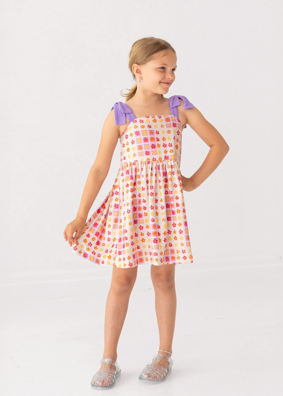 A young girl smiles in the Mabel and Honey Petal Power Tank Sleeve with Knot Detail Dress, featuring a colorful geometric print, purple shoulder knots, and an A-line skirt. She stands on a white background, wearing clear sandals.