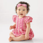 A baby with curly dark hair wears the Mabel and Honey Pinkalicious Short Sleeve Romper and a matching pink headband, sitting barefoot on the floor against a plain light background, smiling and looking to the side.