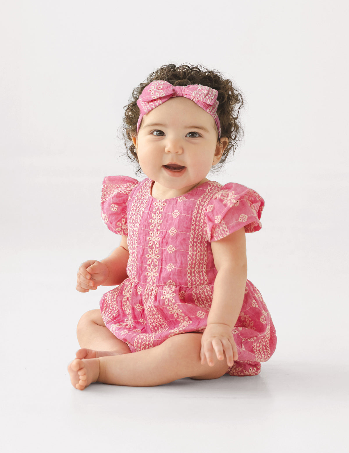 A baby with curly dark hair wears the Mabel and Honey Pinkalicious Short Sleeve Romper and a matching pink headband, sitting barefoot on the floor against a plain light background, smiling and looking to the side.