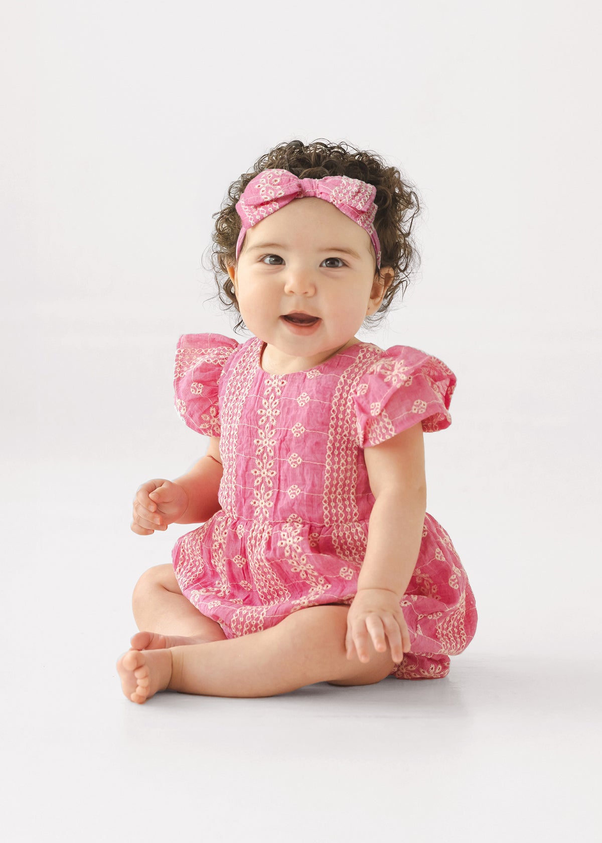 A baby with curly dark hair wears the Mabel and Honey Pinkalicious Short Sleeve Romper and a matching pink headband, sitting barefoot on the floor against a plain light background, smiling and looking to the side.