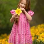 A young girl smiles while holding a yellow daffodil in a blooming field, wearing the Pinkalicious Short Sleeve Dress by Mabel and Honey.