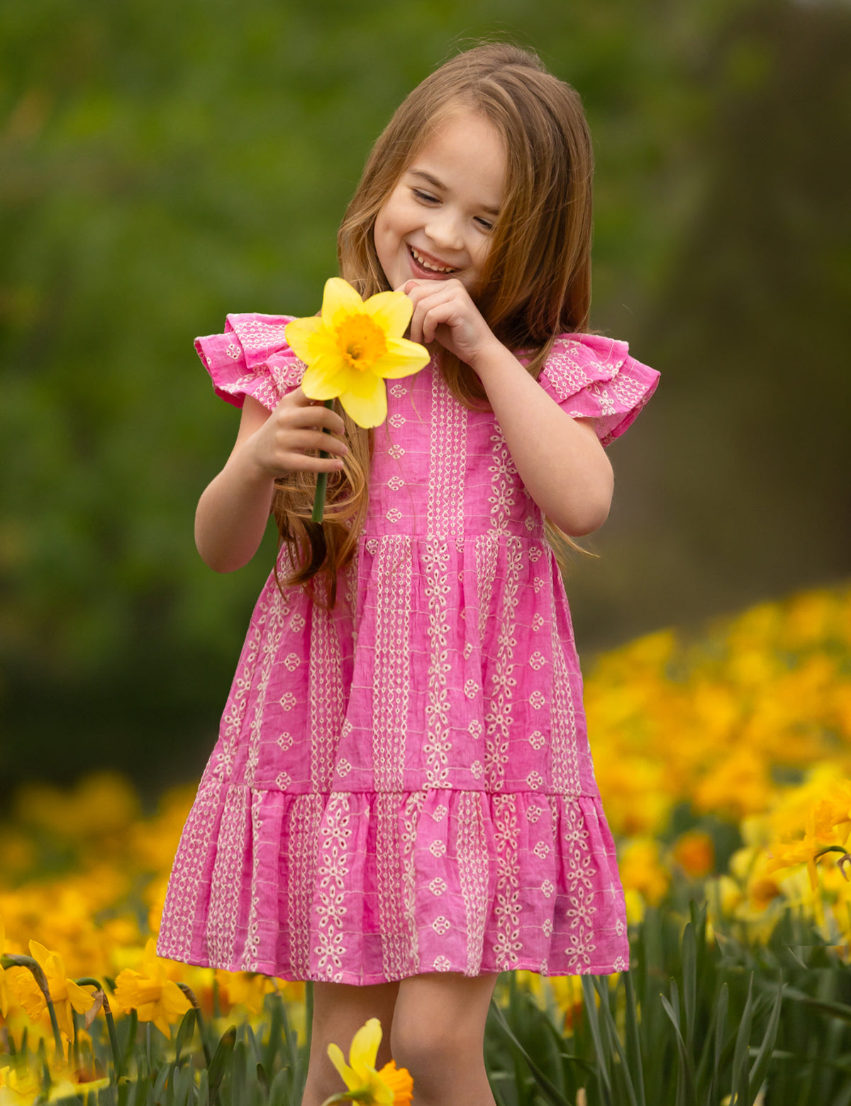 A young girl smiles while holding a yellow daffodil in a blooming field, wearing the Pinkalicious Short Sleeve Dress by Mabel and Honey.