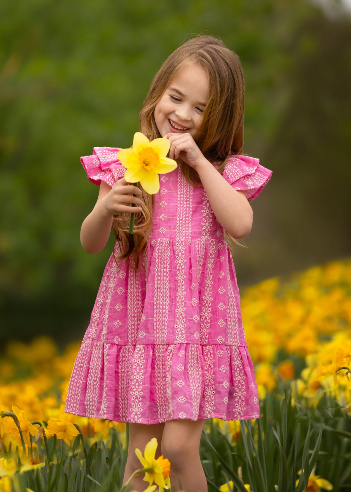 A young girl smiles while holding a yellow daffodil in a blooming field, wearing the Pinkalicious Short Sleeve Dress by Mabel and Honey.