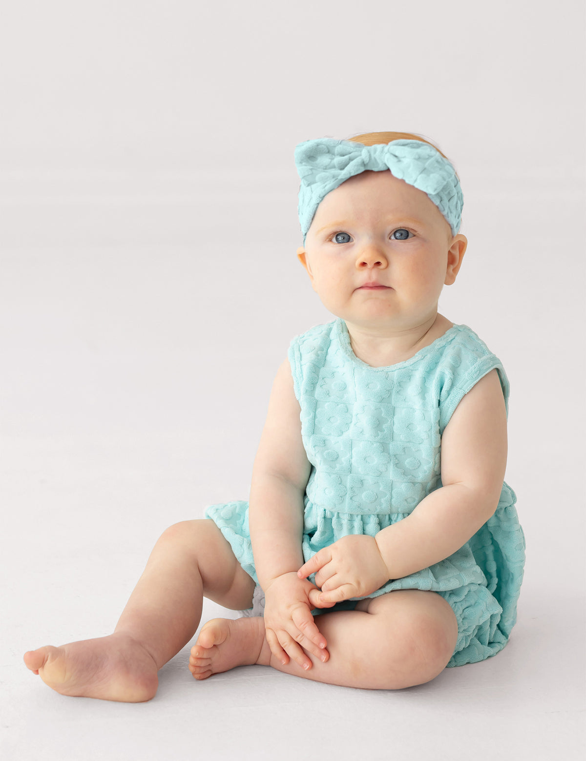 A baby with blue eyes sits on the floor wearing the Mabel and Honey Queenie Sleeveless Romper in light blue terry cloth with a gentle bubble shape and a matching bow headband, gazing slightly upward against a plain white background.
