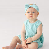 A baby with blue eyes sits on the floor wearing the Mabel and Honey Queenie Sleeveless Romper in light blue terry cloth with a gentle bubble shape and a matching bow headband, gazing slightly upward against a plain white background.