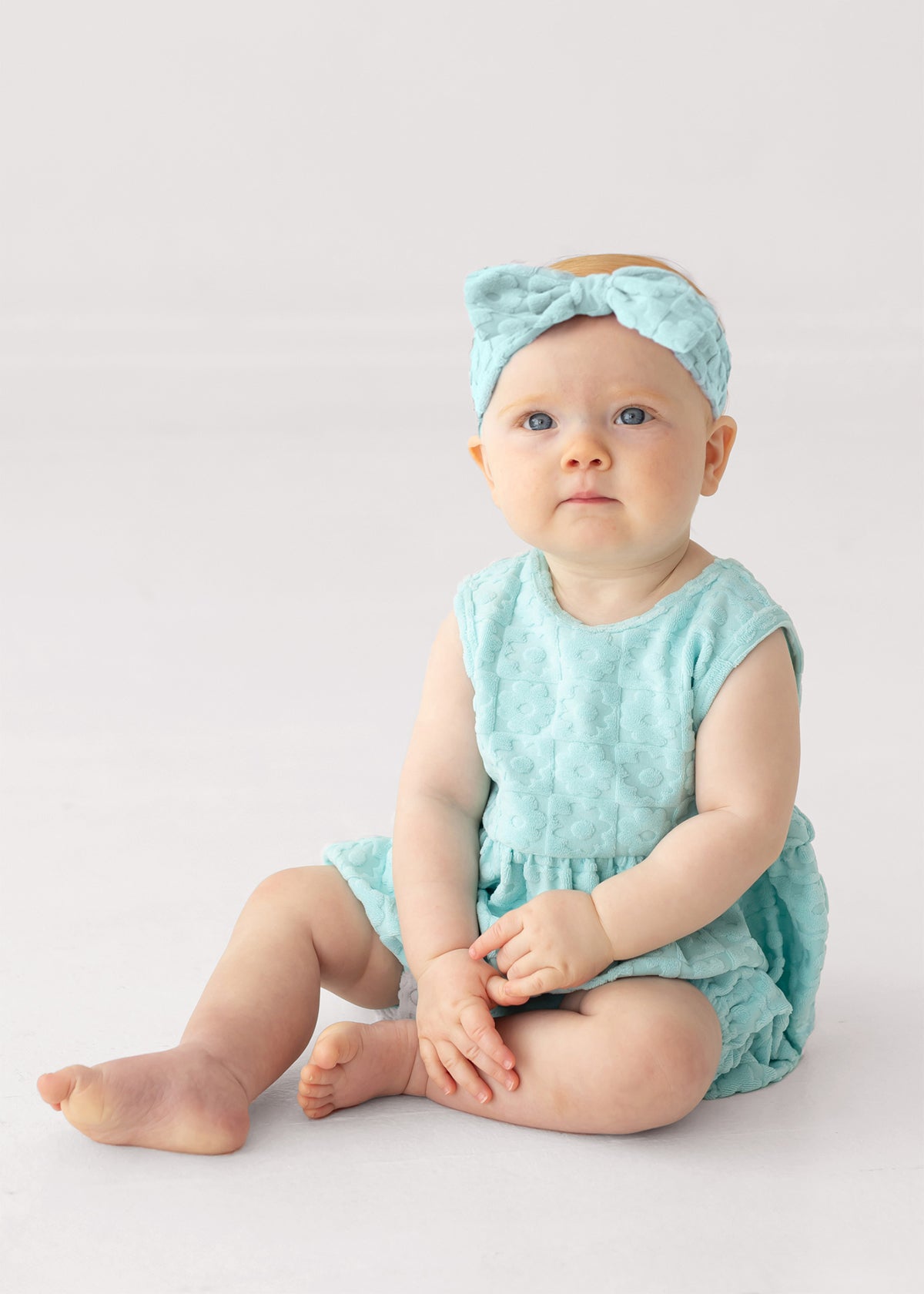 A baby with blue eyes sits on the floor wearing the Mabel and Honey Queenie Sleeveless Romper in light blue terry cloth with a gentle bubble shape and a matching bow headband, gazing slightly upward against a plain white background.
