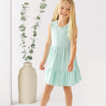 A young girl with long blonde hair smiles in front of a white background, wearing the Mabel and Honey Queenie Sleeveless Dress in light blue and white sandals, beside a tall beige vase with eucalyptus branches.