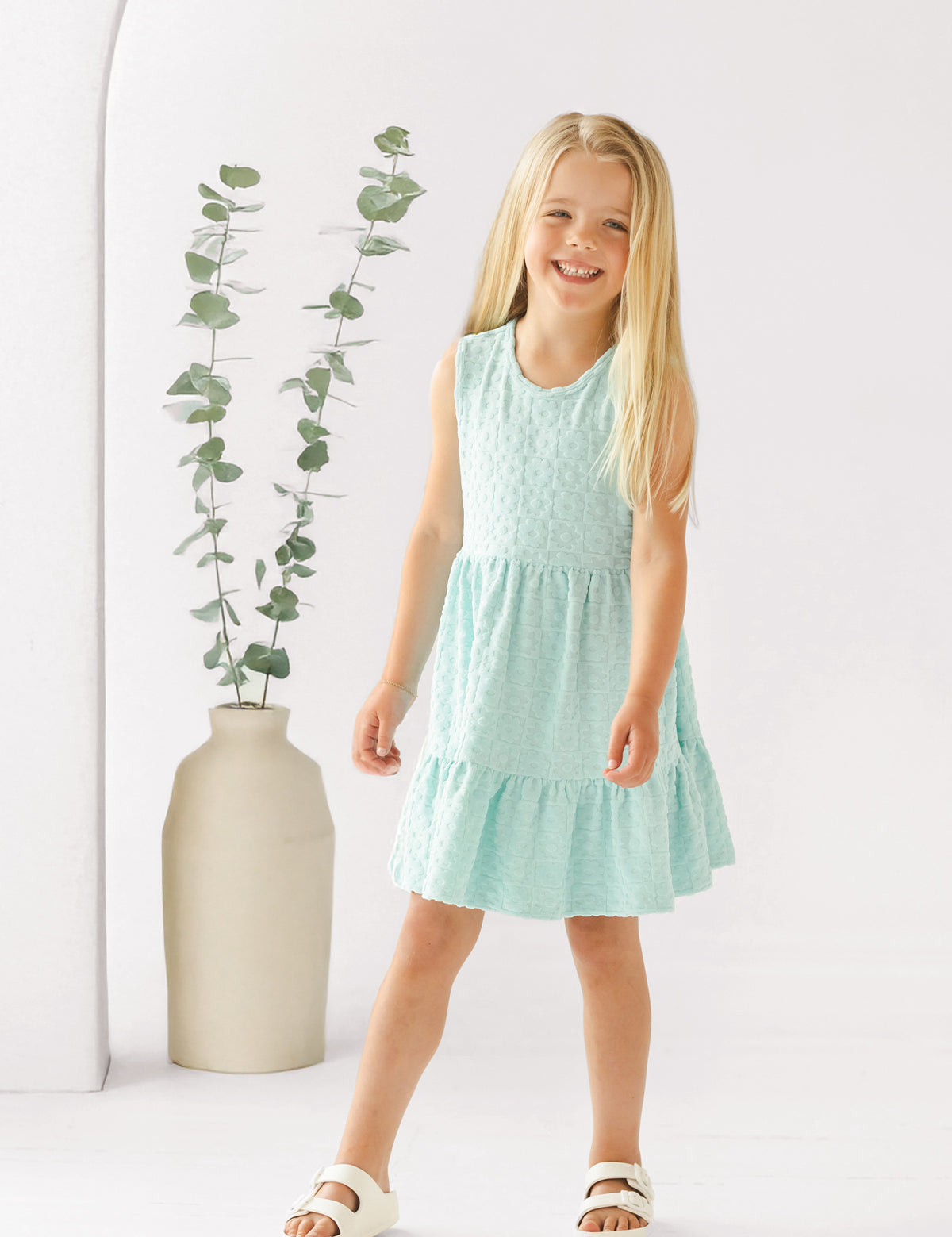 A young girl with long blonde hair smiles in front of a white background, wearing the Mabel and Honey Queenie Sleeveless Dress in light blue and white sandals, beside a tall beige vase with eucalyptus branches.