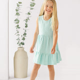 A young girl with long blonde hair smiles in front of a white background, wearing the Mabel and Honey Queenie Sleeveless Dress in light blue and white sandals, beside a tall beige vase with eucalyptus branches.