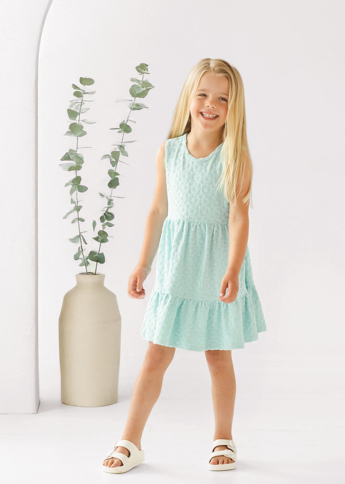 A young girl with long blonde hair smiles in front of a white background, wearing the Mabel and Honey Queenie Sleeveless Dress in light blue and white sandals, beside a tall beige vase with eucalyptus branches.
