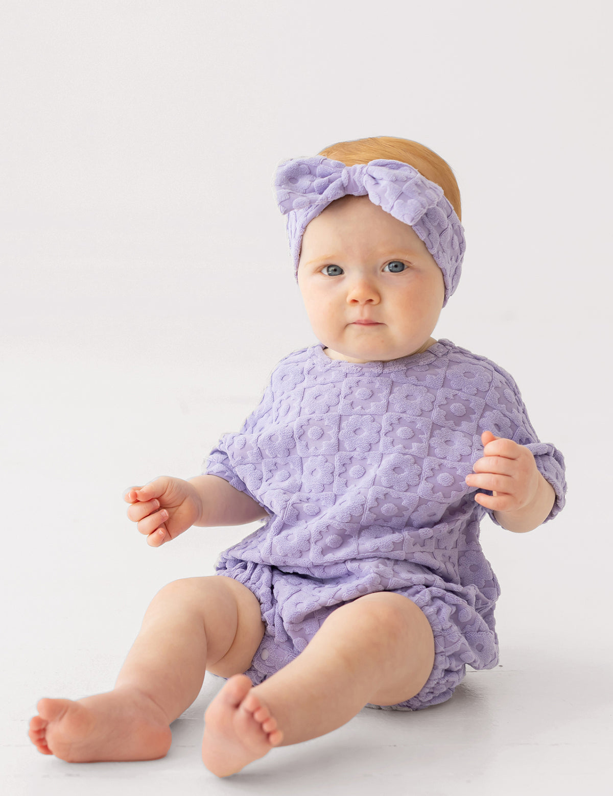 A baby in the Mabel and Honey Veronica Short Sleeve Romper with a matching bow headband sits on a white surface, looking at the camera with a neutral expression.