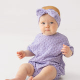 A baby in the Mabel and Honey Veronica Short Sleeve Romper with a matching bow headband sits on a white surface, looking at the camera with a neutral expression.