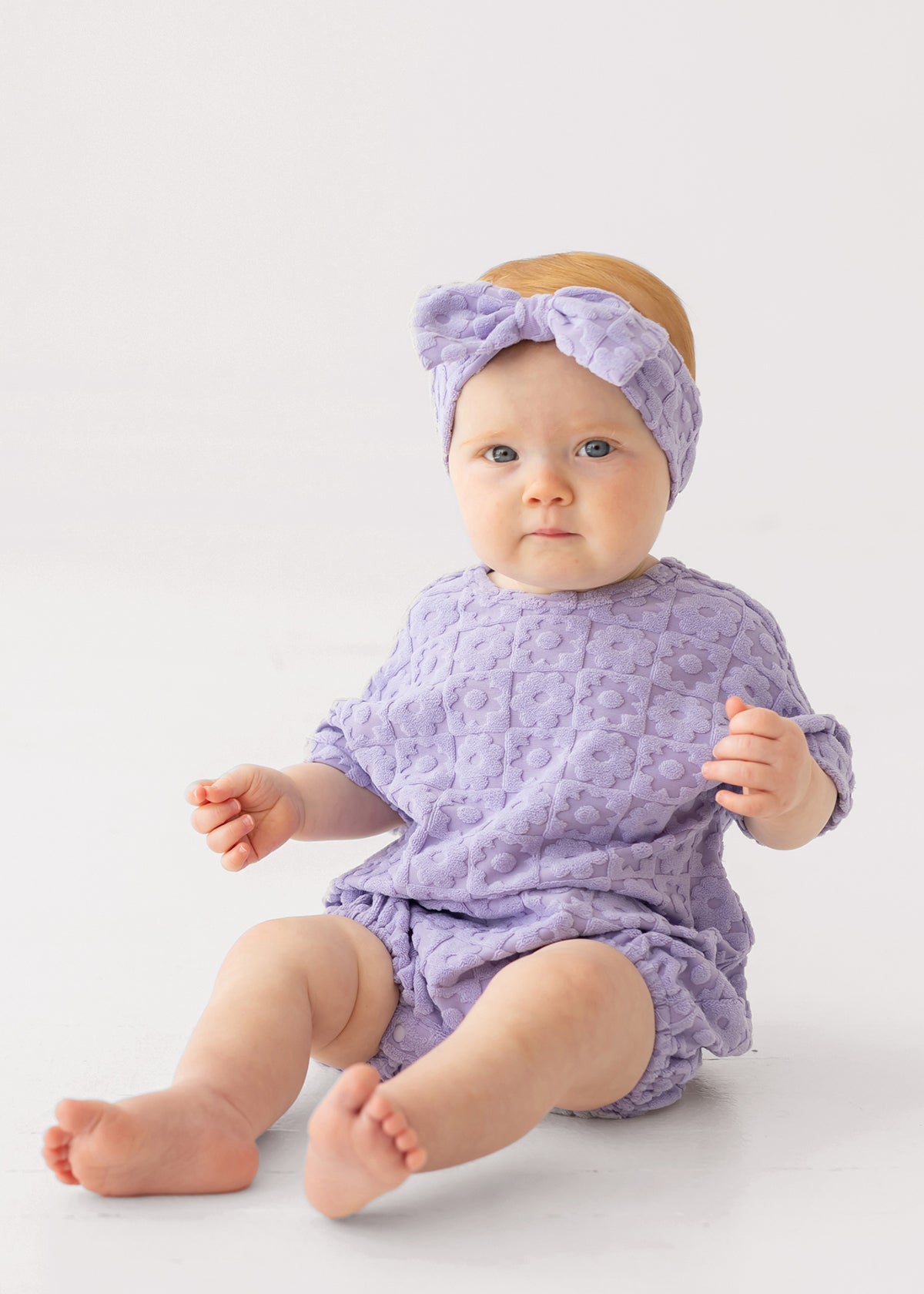 A baby in the Mabel and Honey Veronica Short Sleeve Romper with a matching bow headband sits on a white surface, looking at the camera with a neutral expression.