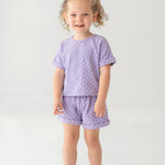 A young child with curly hair smiles while wearing the Mabel and Honey Veronica Short Sleeve Two Piece Set in lavender, featuring elastic waistband shorts. She stands against a plain, light background and wears white sandals.