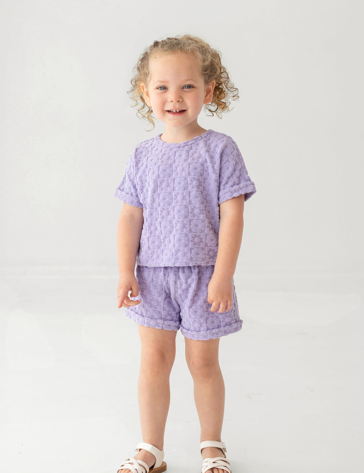 A young child with curly hair smiles while wearing the Mabel and Honey Veronica Short Sleeve Two Piece Set in lavender, featuring elastic waistband shorts. She stands against a plain, light background and wears white sandals.