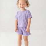 A young child with curly hair smiles while wearing the Mabel and Honey Veronica Short Sleeve Two Piece Set in lavender, featuring elastic waistband shorts. She stands against a plain, light background and wears white sandals.