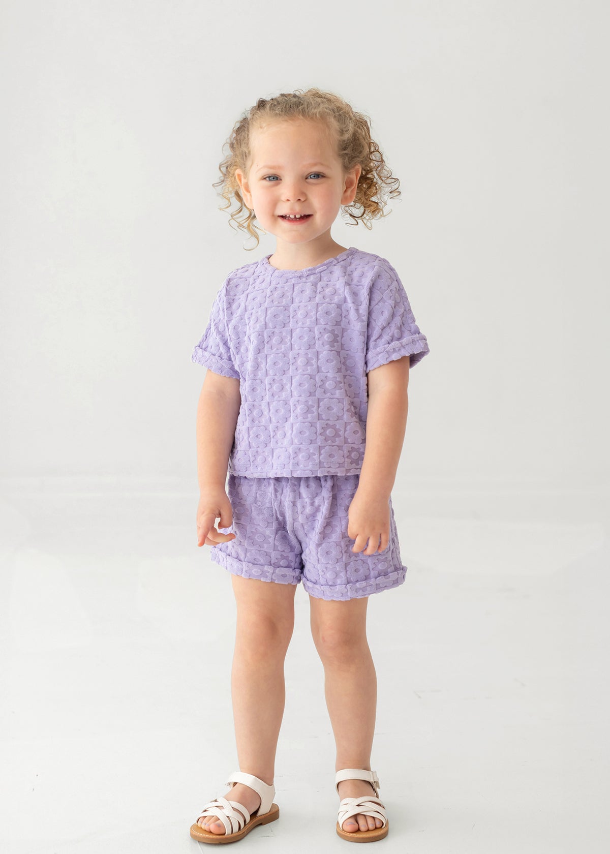 A young child with curly hair smiles while wearing the Mabel and Honey Veronica Short Sleeve Two Piece Set in lavender, featuring elastic waistband shorts. She stands against a plain, light background and wears white sandals.