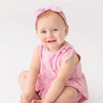A smiling baby with light skin, blue eyes, and light hair sits barefoot on a white background, wearing Mabel and Honey's Tilly Sleeveless with Knot Detail Romper in pink and a matching bow headband.