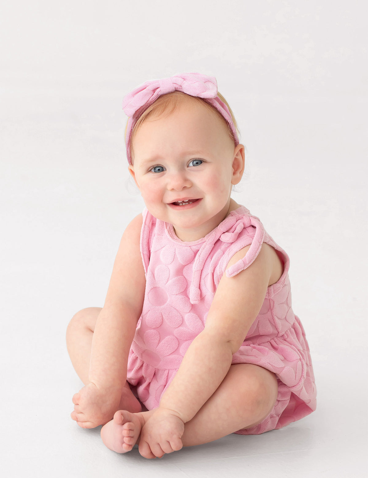A smiling baby with light skin, blue eyes, and light hair sits barefoot on a white background, wearing Mabel and Honey's Tilly Sleeveless with Knot Detail Romper in pink and a matching bow headband.
