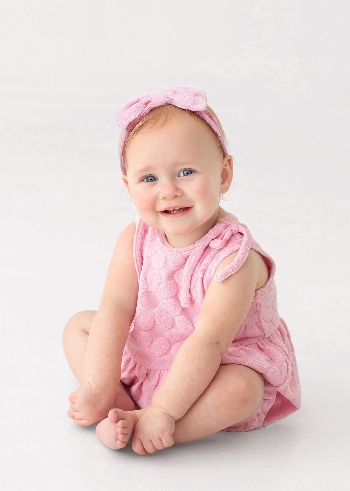 A smiling baby with light skin, blue eyes, and light hair sits barefoot on a white background, wearing Mabel and Honey's Tilly Sleeveless with Knot Detail Romper in pink and a matching bow headband.