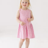 A young child with blonde hair smiles against a white background, wearing the Mabel and Honey Tilly Short Sleeve Dress and white sneakers.