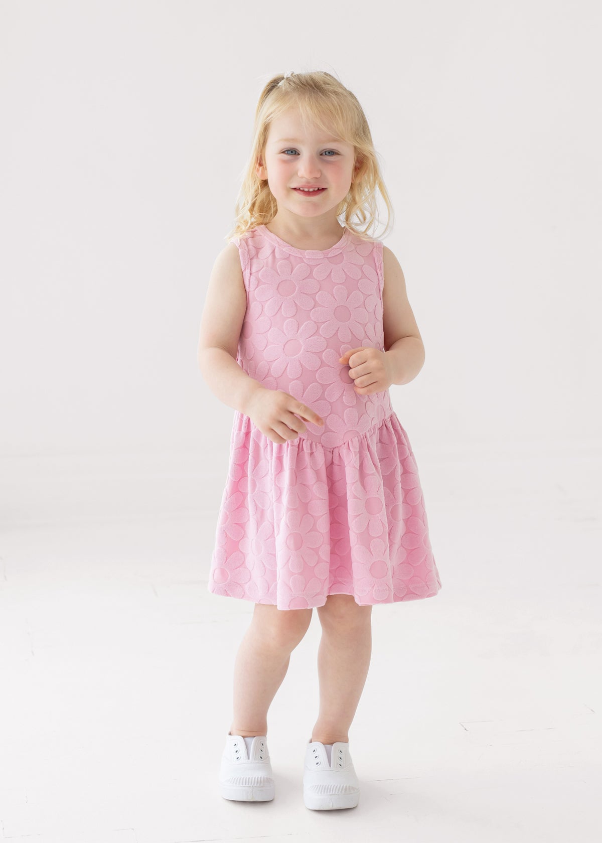 A young child with blonde hair smiles against a white background, wearing the Mabel and Honey Tilly Short Sleeve Dress and white sneakers.