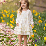 A young girl wears the Mabel and Honey Sweet Cottontails Sleeveless Dress, smiling among yellow daffodils and pink petals with flowering trees in the background on a sunny day.