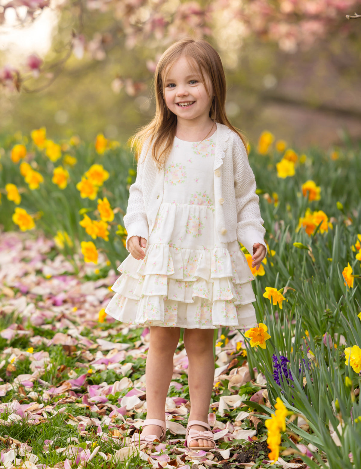A young girl wears the Mabel and Honey Sweet Cottontails Sleeveless Dress, smiling among yellow daffodils and pink petals with flowering trees in the background on a sunny day.