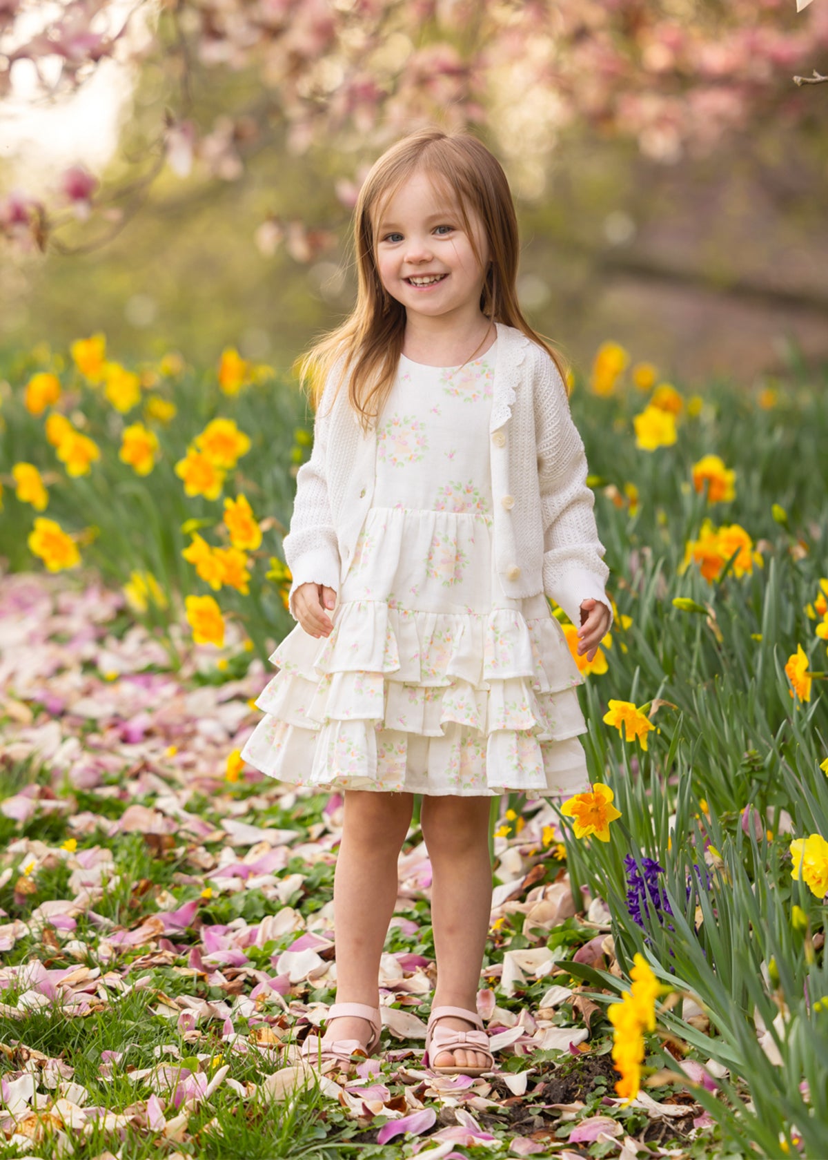 A young girl wears the Mabel and Honey Sweet Cottontails Sleeveless Dress, smiling among yellow daffodils and pink petals with flowering trees in the background on a sunny day.